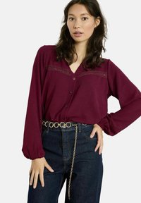 Blusa bordeaux a maniche lunghe con chiusura a bottoni, dettagli in pizzo sulle spalle e vestibilità rilassata, abbinata a jeans scuri a vita alta e cintura a catena.