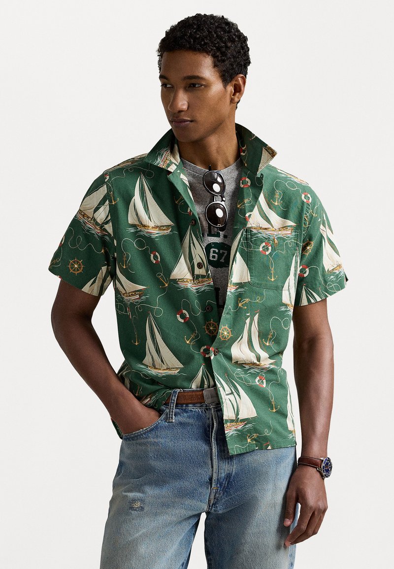 Korte mouw groene shirt met een zeilmotief, witte zeilen, rode zwemringen en marineblauwe ankers. Draagt over een grijs T-shirt en jeans.