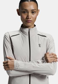 Chaqueta de running gris claro con un cuello alto, cremallera negra y sutiles acentos negros en los hombros. Textura suave y diseño ajustado.