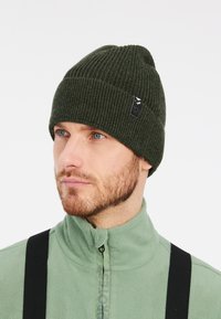 Beanie a coste verde scuro con orlo ripiegato e una piccola etichetta nera del marchio. Indossato con una giacca in fleece verde chiaro.