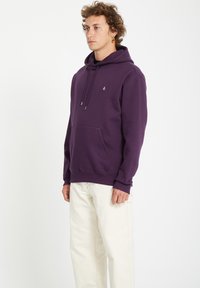 Lilla hoodie lavet af blødt stof, med en kængurulomme, justerbar snoretræk ved hætten og et lille logo på brystet.