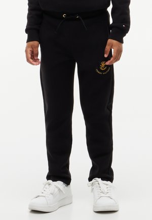 Person iført sorte Tommy Hilfiger joggingbukser med guldlogo og hvide sneakers, står med hænderne afslappet langs siderne.