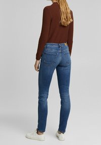 Blå denim slim fit jeans med midjehöjd, bakfickor och subtila sömnadsdetaljer, i kombination med en brun texturerad tröja och vita sneakers.