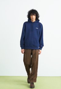 Navy blue hooded sweatshirt with a front pocket and drawstring, paired with brown wide-leg trousers and dark shoes, standing on green flooring.