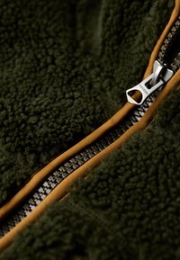 Dunkelgrüner Fleece-Stoff mit strukturierter Oberfläche, akzentuiert mit einem kontrastierenden Reißverschlussbesatz in Senfgelb und silberner Reißverschlussbeschlägen.
