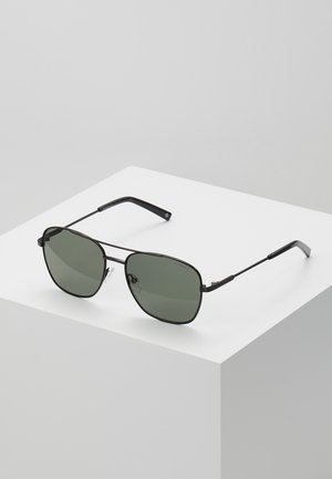 Occhiali da sole in stile black metal con lenti verdi, forma ottagonale, aste sottili e dettagli texturizzati sulla montatura.