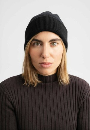 Schwarze gerippte Strickmütze mit passendem Design, getragen von einer Person mit schulterlangem blonden Haar und einem dunklen gerippten Pullover.