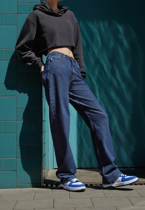 Personne portant un sweat à capuche noir court, un jean bleu ample et des baskets blanches et bleues, debout contre un mur carrelé turquoise au soleil.