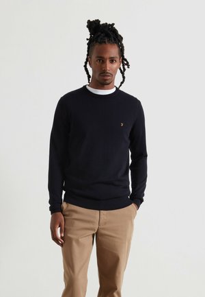 Marineblauwe sweater met ronde hals, gemaakt van een gladde breiwerkstof, met een klein gouden logo op de linkerkant van de borst. Gelijkend op beige broek.