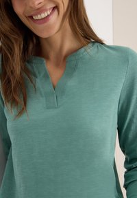 Haut turquoise à manches longues avec un décolleté en V et des détails cousus. Texture lisse avec une coupe décontractée. Fond de couleur neutre.