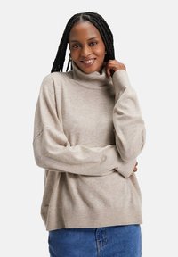 Pull en col roulé beige avec une coupe ample et des poignets côtelés, fabriqué en matériau doux. Porté par-dessus un vêtement bleu, il présente un accent de logo.