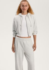 Svetlošedá zipová mikina s kapucňou a šnúrkami, v kombinácii s rovnakými svetlošedými elastickými nohavicami, oboje vyrobené z mäkkej látky.