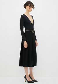 Bodysuit negro de manga larga con un profundo escote en V, combinado con una falda negra de talle alto y pliegues y tacones negros de punta. Detalle de cinturón con hebilla.