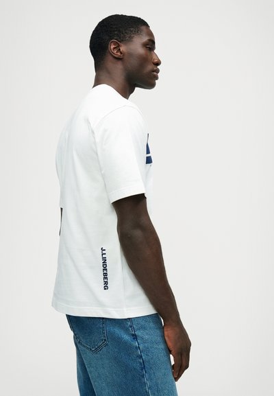 Baldasis medvilninis marškinėlis su mėlynu grafiniu dizainu priekyje ir "J.LINDEBERG" tekstu šone. Trumpomis rankovėmis, laisvas siluetas.