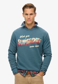 Sudadera verde azulado con el gráfico "Wish you SUPERDRY were here". Manga larga, cuello acanalado. Capas con una camisa de cuadros debajo.
