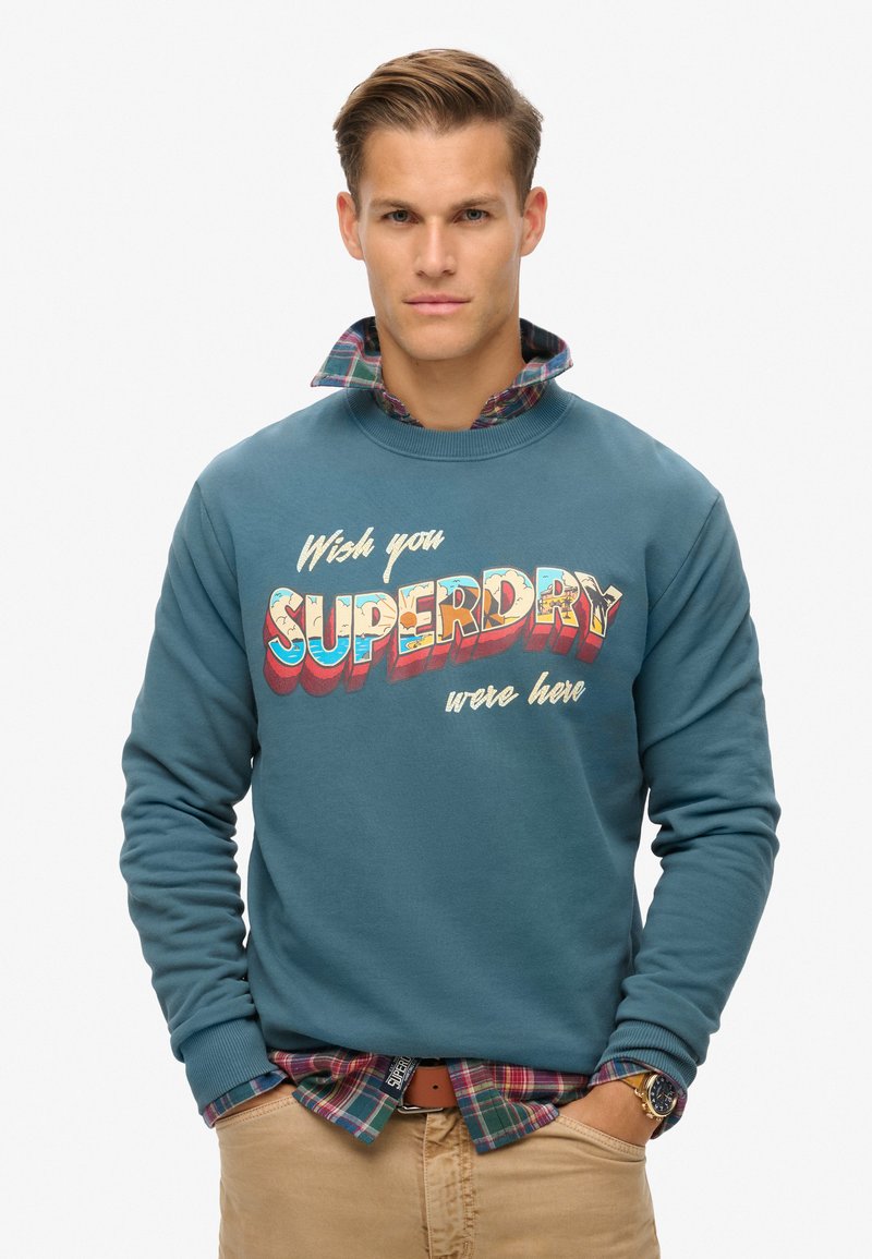 Sudadera verde azulado con el gráfico "Wish you SUPERDRY were here". Manga larga, cuello acanalado. Capas con una camisa de cuadros debajo.
