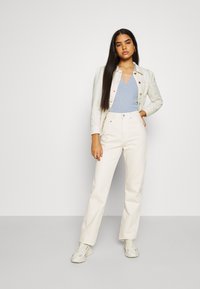 Giacca di jeans bianca e pantaloni, top azzurro chiaro con scollatura a V e scarpe da ginnastica chiare. Caratteristiche includono una chiusura frontale con bottoni e un taglio dritto.