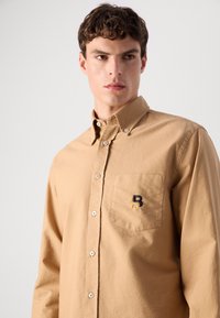 Langermet skjorte med knapper i khaki bomull, med en brystlomme med en mørk og lys brodert logo. Klassisk kragedesign.