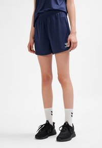 Mujer con pantalones cortos deportivos azul marino, calcetas blancas hasta la mitad de la pantorrilla con chevrones negros, y zapatos deportivos negros, de pie sobre un fondo blanco.