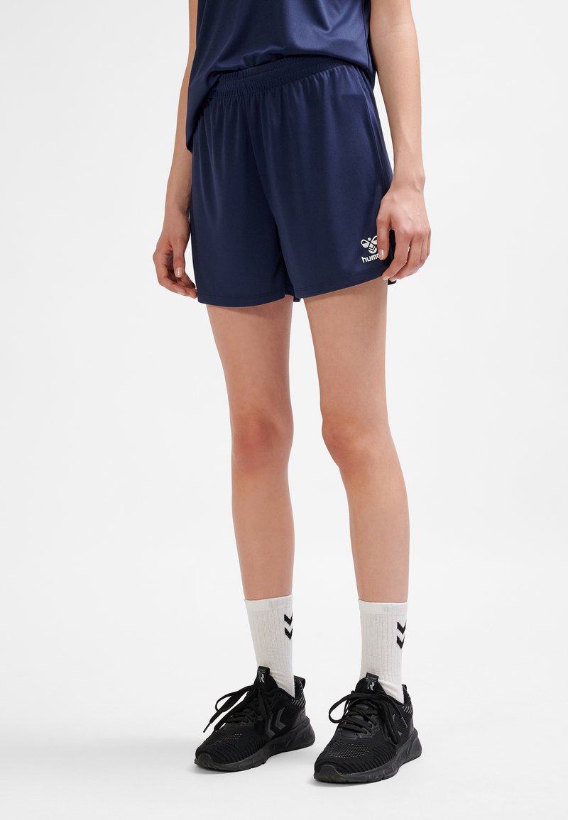 Mujer con pantalones cortos deportivos azul marino, calcetas blancas hasta la mitad de la pantorrilla con chevrones negros, y zapatos deportivos negros, de pie sobre un fondo blanco.