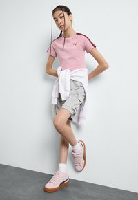 Chemise rose à manches courtes avec des accents bordeaux, shorts cargo gris et baskets roses avec des semelles marron et des chaussettes blanches. Pull blanc noué autour de la taille.