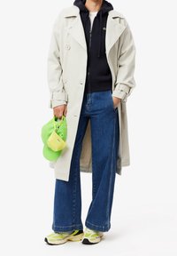 Lichtgrijze trenchcoat over een zwarte hoodie, blauwe wijde jeans, groene pet en kleine gele tas. Sneakers met neon accenten en zwarte zolen.