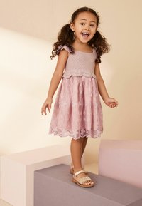 Rosa Blumenkleid mit gehäkeltem Oberteil und Rüschensäulen. Verfügt über bestickte Blumen und einen gewellten Saum. Kombiniert mit verzierten Sandalen mit Riemen.