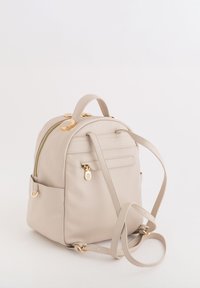 Beżowy plecak ze skórzanym wykończeniem, ze złotymi akcentami, zamknięciem na zamek, górną rączką, regulowanymi paskami i boczną kieszenią na dodatkową przestrzeń do przechowywania.