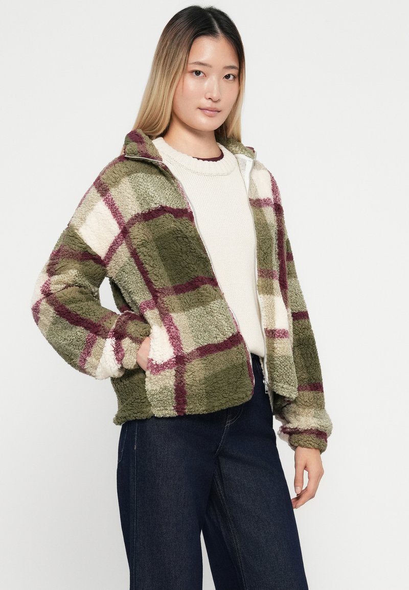 Giacca in pile con motivo a quadri verde, avorio e bordeaux. Dotata di zip frontale e due tasche laterali. Si abbina a un maglione di maglia color crema indossato sotto.