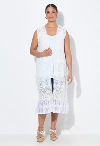 Gilet in pizzo bianco con apertura frontale, presenta motivi intricati e un orlo arricciato. Indossato sopra una maglietta bianca, abbinato a sandali espadrillas.