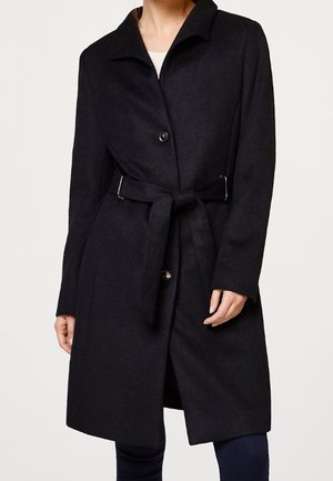 Femme portant un manteau noir ceinturé longueur genou avec col et deux boutons, sur un pantalon foncé, posant devant un fond clair uni.