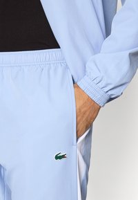 Άνδρας που φοράει ανοιχτό μπλε αθλητικό παντελόνι και σακάκι με το χέρι στην τσέπη, με πράσινο λογότυπο κροκοδείλου στο παντελόνι.