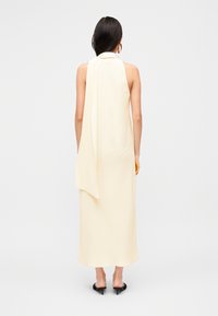 Μακρύ κρεμ φόρεμα με halter λαιμόκοψη και ρέουσα κορδέλα στο πίσω μέρος. Το ύφασμα είναι λείο, με ευθεία σιλουέτα, συνδυασμένο με μαύρα πέδιλα ανοιχτού δαχτύλου.