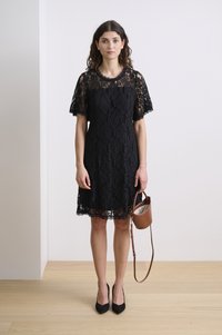 Black lace dress with a round neckline, short flutter sleeves, and scalloped hem. Paired with a small brown handbag and black heels.