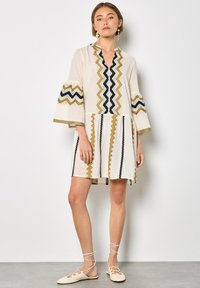 Robe crème à manches courtes avec des motifs en zigzag noirs et dorés, dotée d'un décolleté en V et de manches évasées. Tissu léger, longueur au genou.