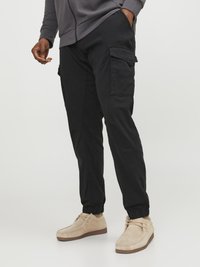 Zwarte cargobroek met elastische boorden, voorzien van meerdere zijzakken en een relaxte pasvorm, gecombineerd met lichtbeige suède schoenen.