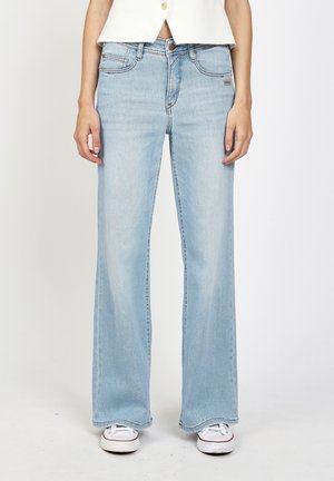 Hellblaue Jeans mit hoher Taille und weitem Bein, kombiniert mit weißen Turnschuhen und einem weißen bauchfreien Oberteil, dargestellt von der Taille bis zu den Füßen.