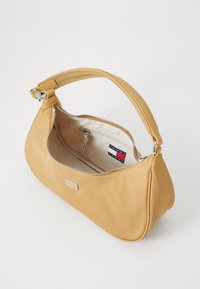 Žuta najlonska torbica s zakrivljenim oblikom, srebrnim patentnim zatvaračem i kratkom drškom. Unutrašnjost sadrži mali zip džep i Tommy Hilfiger brendiranje.