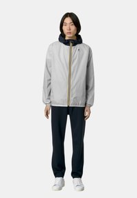 Giacca grigia chiara con cappuccio blu navy, zip frontale e strisce gialle; abbinata a pantaloni scuri e sneakers bianche.