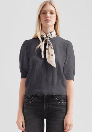 Junge Frau mit langen blonden Haaren trägt übergroße Brille, einen dunklen Kurzarmpullover, schwarze Jeans mit Metallösen und ein weiß gemustertes Halstuch.