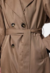 Brun trenchcoat med en dobbeltradet design, med svarte knapper, et knyttet midjebelte og sidelommer. Glatt, teksturert stoff.