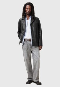 Schwarze Lederjacke mit Knopfverschluss und zwei Taschen, über einem hellblauen Hemd getragen, kombiniert mit ausgewaschenen grauen Jeans und schwarzen Schuhen.