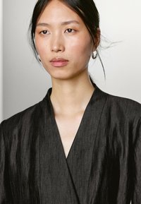 Zwarte textuur kleding met een V-hals ontwerp en een zachte, vloeiende stof. Model heeft oorbellen met een ring en een egale huidskleur.