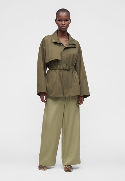 Μοντέλο φορά μια oversized ελιάς πράσινη ζώνη σακάκι και φαρδιά ανοιχτοπράσινα παντελόνια με ανοιχτά σανδάλια, στέκεται μπροστά σε ένα απλό φόντο.