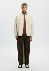 Beige Hemd mit Knöpfen über einem braunen Reißverschlussoberteil, kombiniert mit dunkelbraunen weit geschnittenen Hosen und schwarz-weißen Schuhen.
