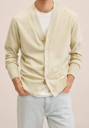 Mann trägt eine cremefarbene, geknöpfte Strickjacke über einem weißen T-Shirt mit hellblauen Jeans, die Hände in den Taschen, vor neutralem Hintergrund.