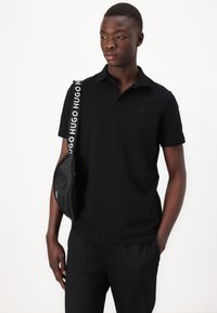 Black polo shirt with a subtle logo, paired with black pants. A black bag with a logo strap is worn across the shoulder.