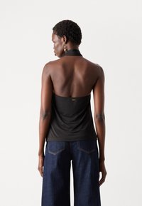 Black halter top made of smooth fabric, featuring a scooped back, and a gold logo detail at the center. Worn with wide-leg dark denim jeans.