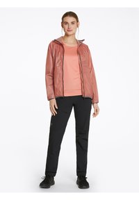 Chaqueta ligera de color rosa, con capucha y cierre frontal, llevada sobre una blusa ajustada melocotón y unos pantalones negros ajustados con bolsillos con cremallera y zapatos negros.