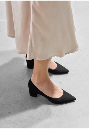 Black suede block-heeled shoes with a pointed toe and a low-cut vamp, worn with a smooth blush-colored skirt. Concrete floor background.